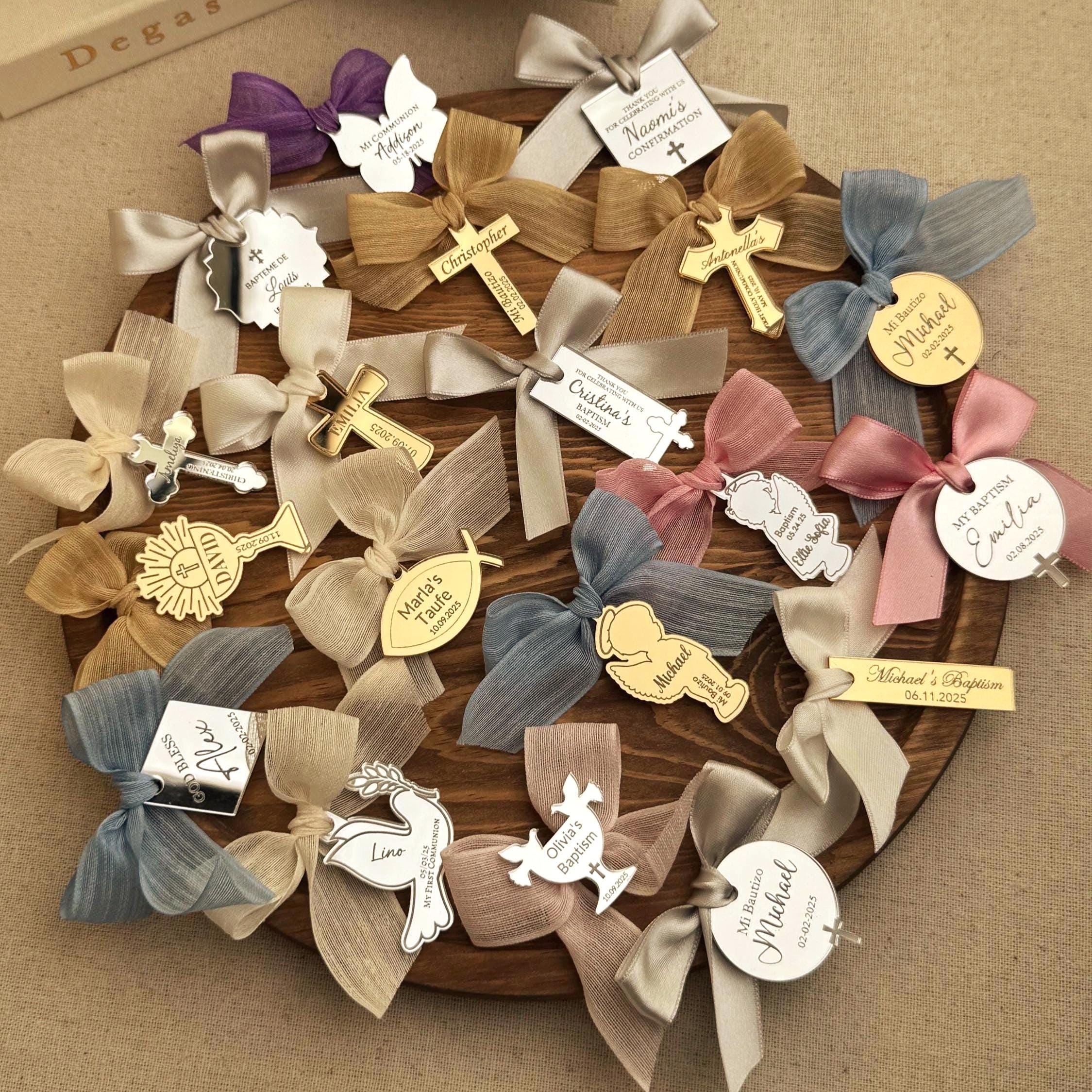 May include: A collection of small, personalized, gold and silver mirror favors with bows. The favors feature various religious symbols, including crosses, doves, angels, and chalices. The favors are decorated with names, dates, and messages, such as 'My Baptism', 'Naomi's Confirmation', 'Cristina's Baptism', and 'Michael's Baptism'. The favors are arranged on a wooden surface.